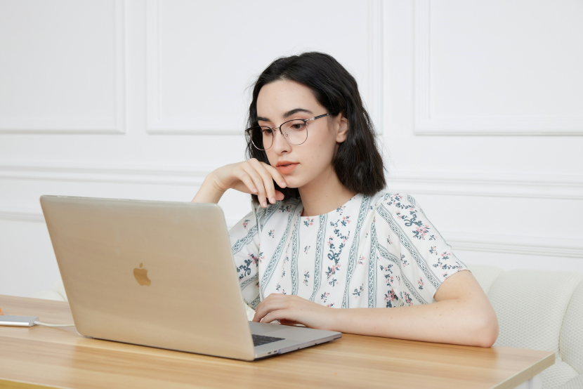 眼鏡をかけた若い女性がノートパソコンを見つめながら机に向かって作業している様子。白い背景の室内で集中している表情。
